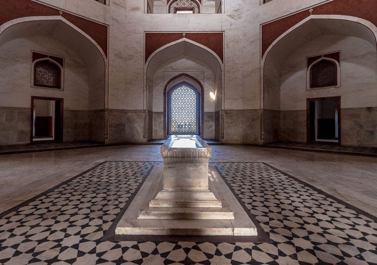 The interior of a large, empty room in a historical building, Humayun's tomb, with a high ceiling, arches, a large window, and a stone structure